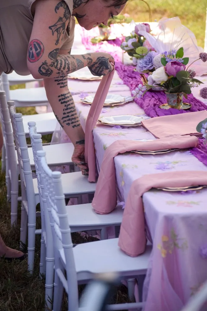 Event planner adjusting a dusty-rose napkin over a lavender gold-rimmed place setting