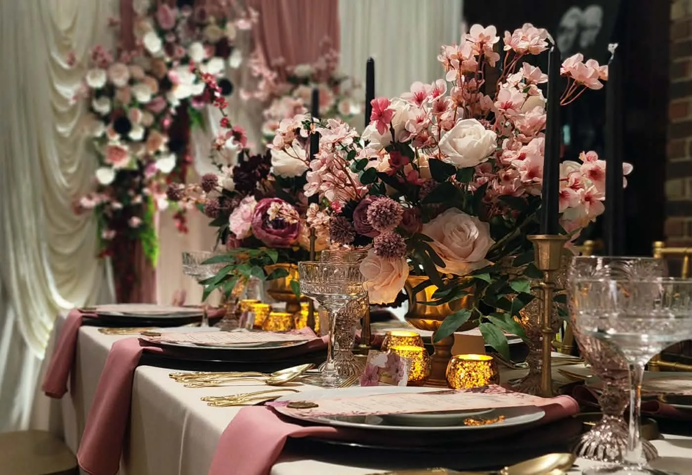 Long dinner table with cherry blossom centerpieces, black taper candles, and blush napkins before a floral draped backdrop